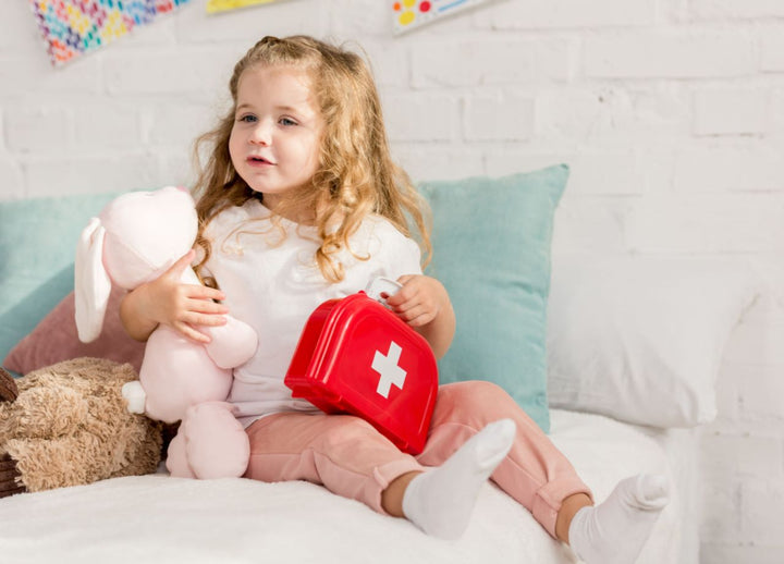 Little girl sitting in bed holding a stuffed teddy and a red plastic first aid bag