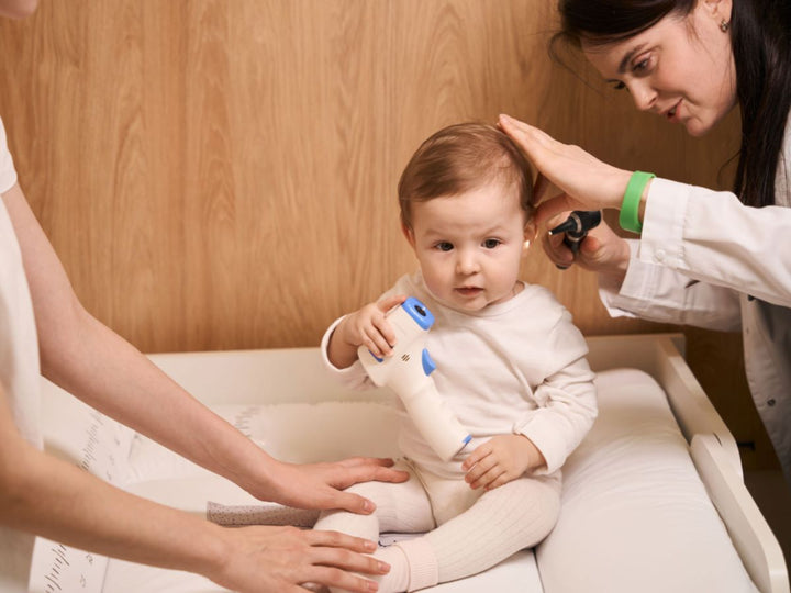 A baby sitting on a bed while the dr inspects her ear