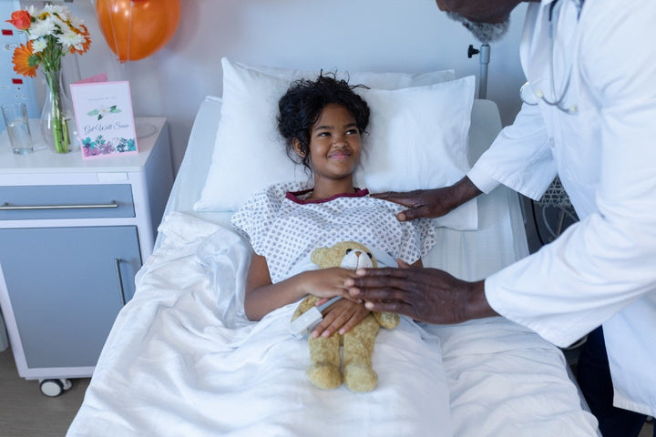  male doctor reassuring smiling sick girl in hospital bed holding teddy