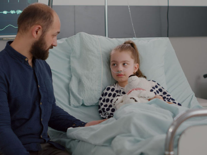 A child in a hospital bed with her father sitting beside her