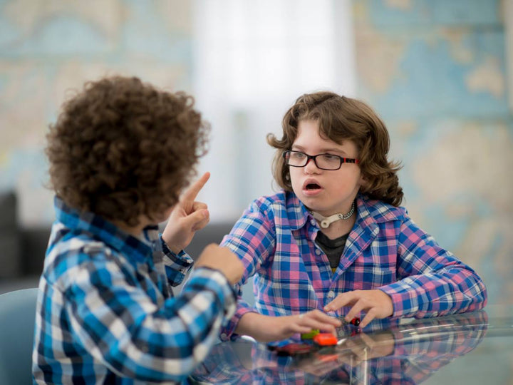 Two children sit on a table and talk, one has a motor speech Impairment device on his throat