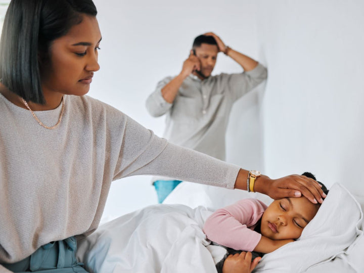 A father holding his head whilst on the phone. The mother is beside the child feeling her temperature with her palm.